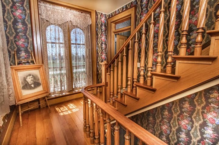 Vintage interior with wooden staircase, patterned wallpaper, and a framed picture resting on the floor.