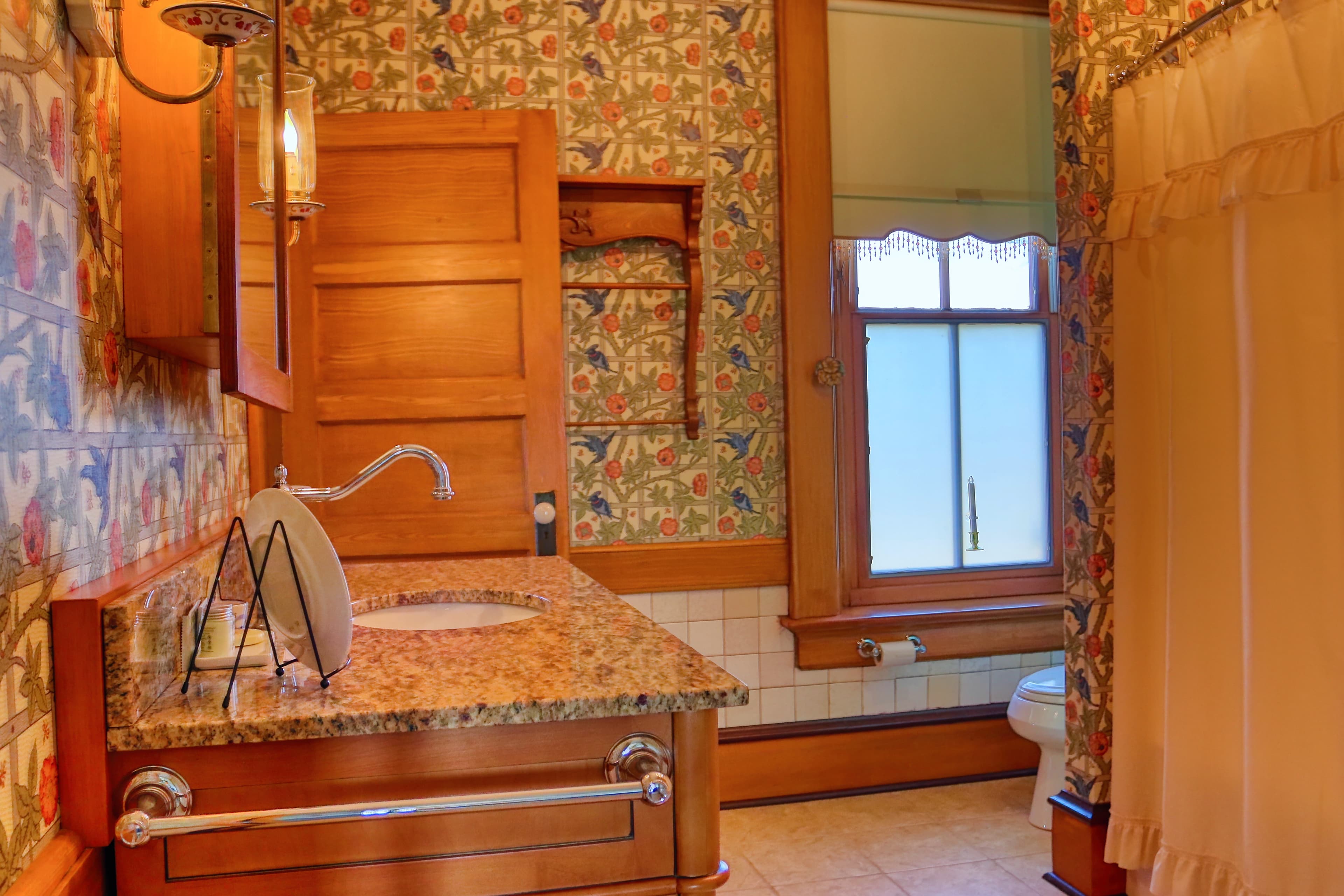 A bathroom with floral wallpaper, shower stall, toilet, and vanity with granite countertop.