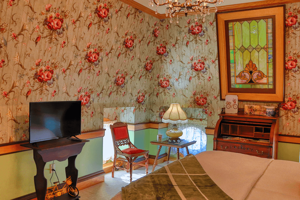 Cozy bedroom featuring floral wallpaper, a TV, antique furniture, and a stained-glass window.