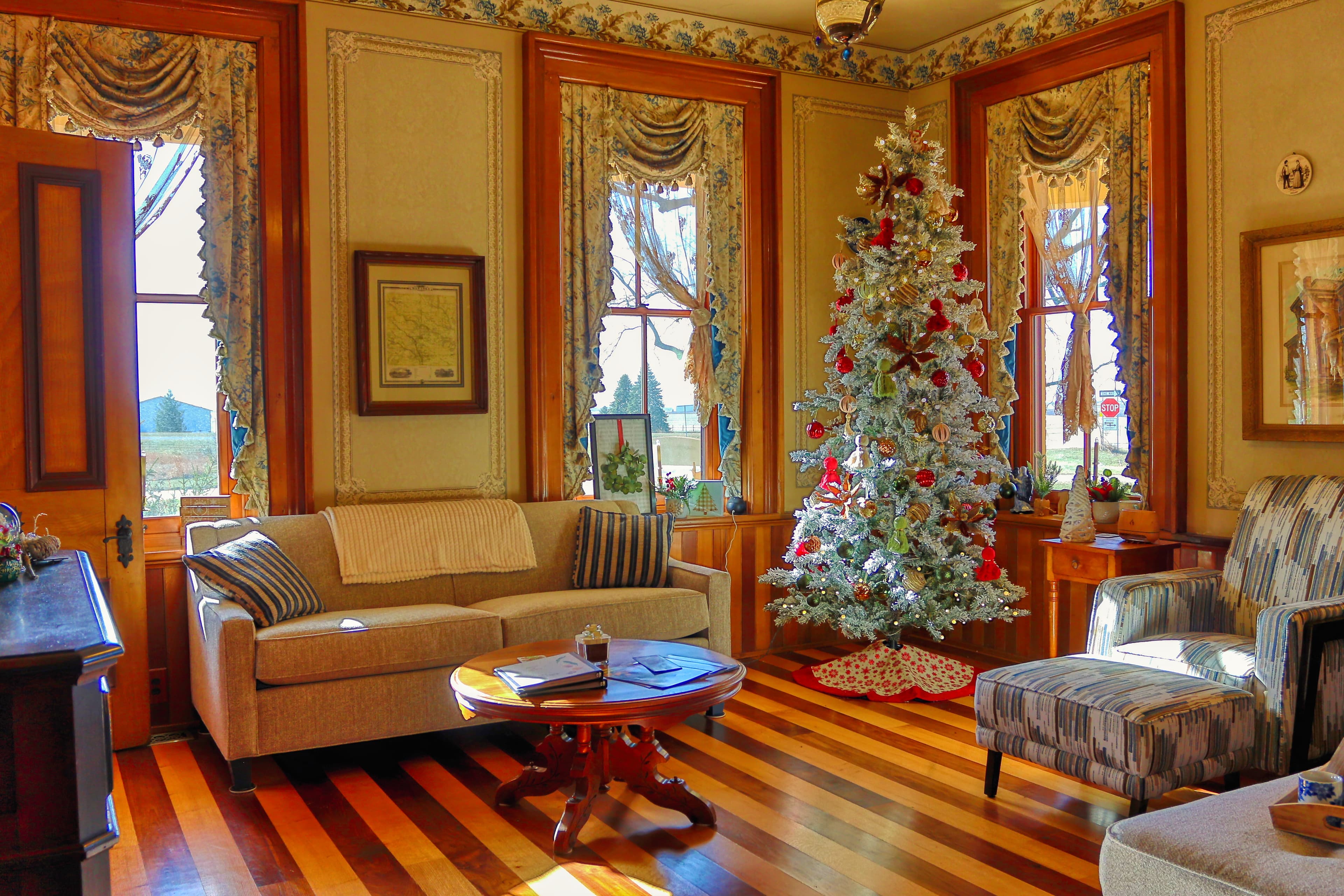 A cozy living room featuring a decorated Christmas tree, a plush sofa, and large windows with floral curtains.