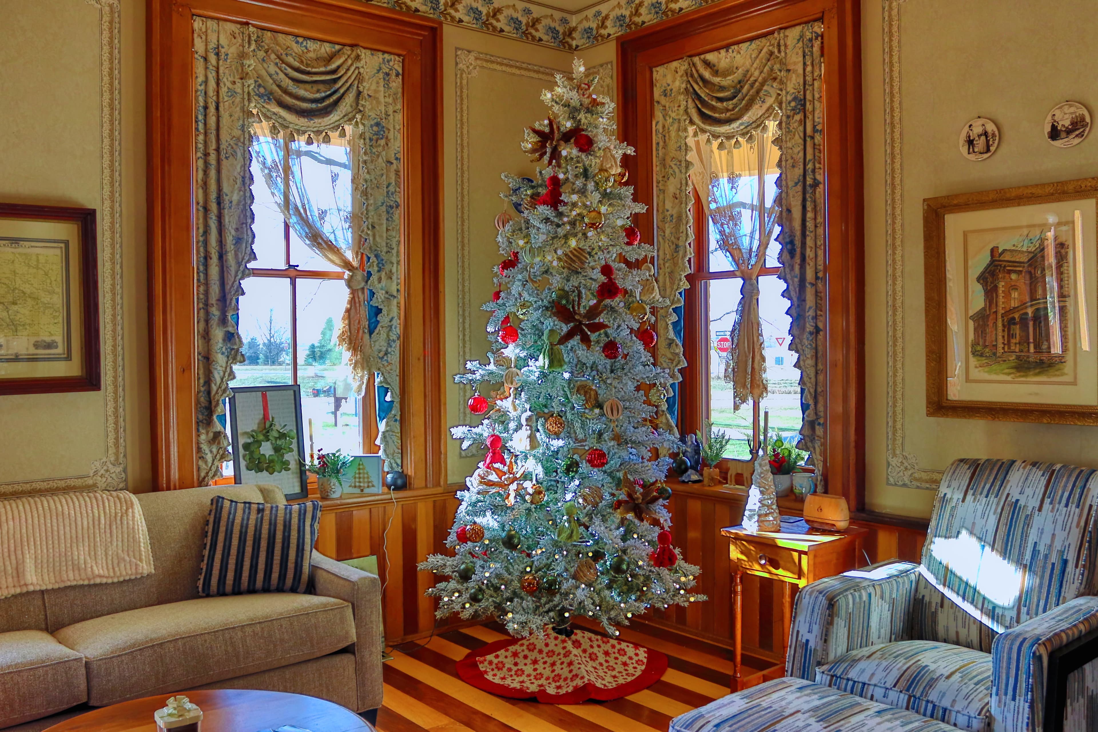 A decorated white Christmas tree stands in a cozy room with vintage furnishings and large windows.