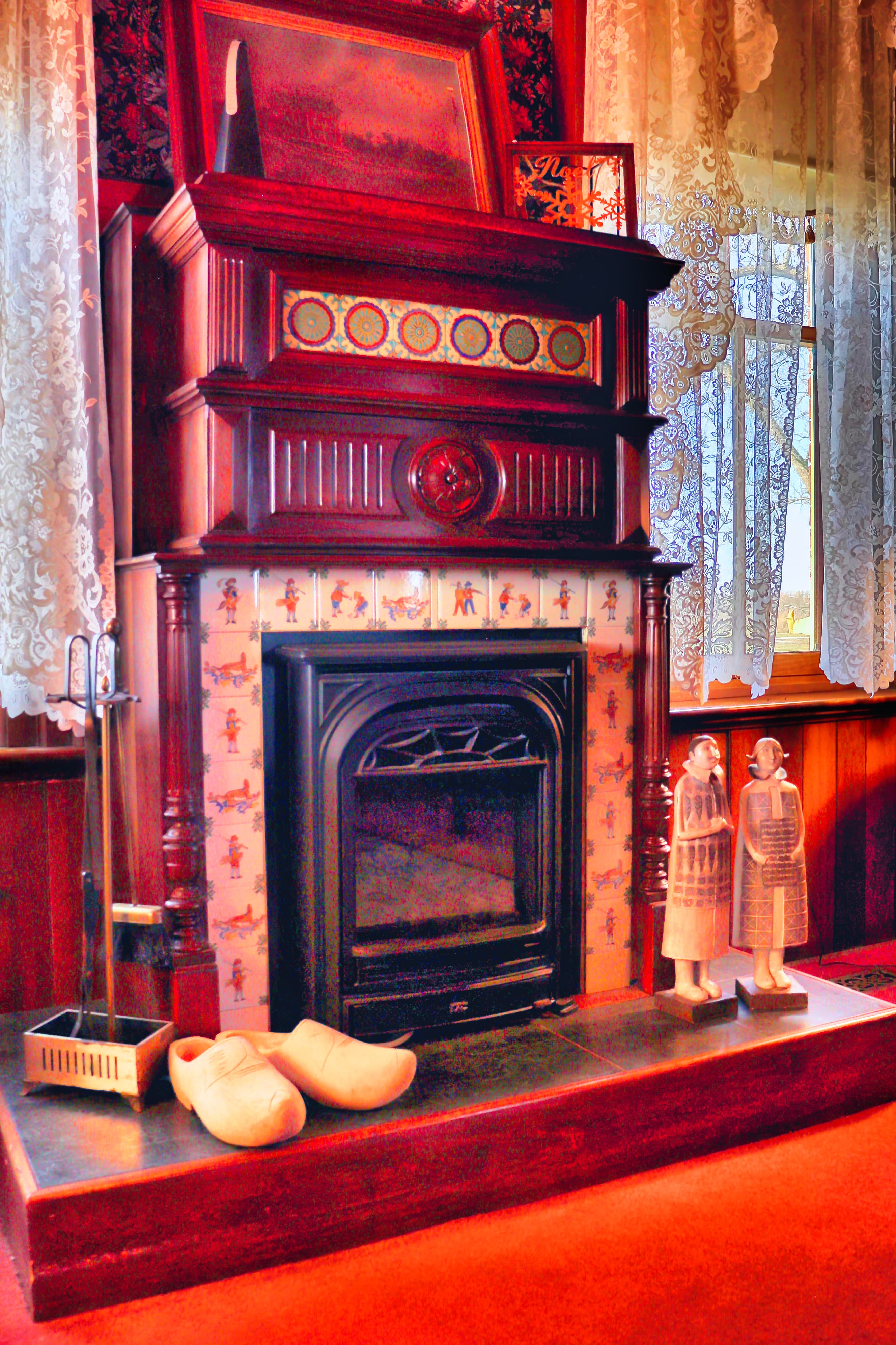 A decorative fireplace with a wooden mantel, two statues, and wooden clogs on a red carpet.