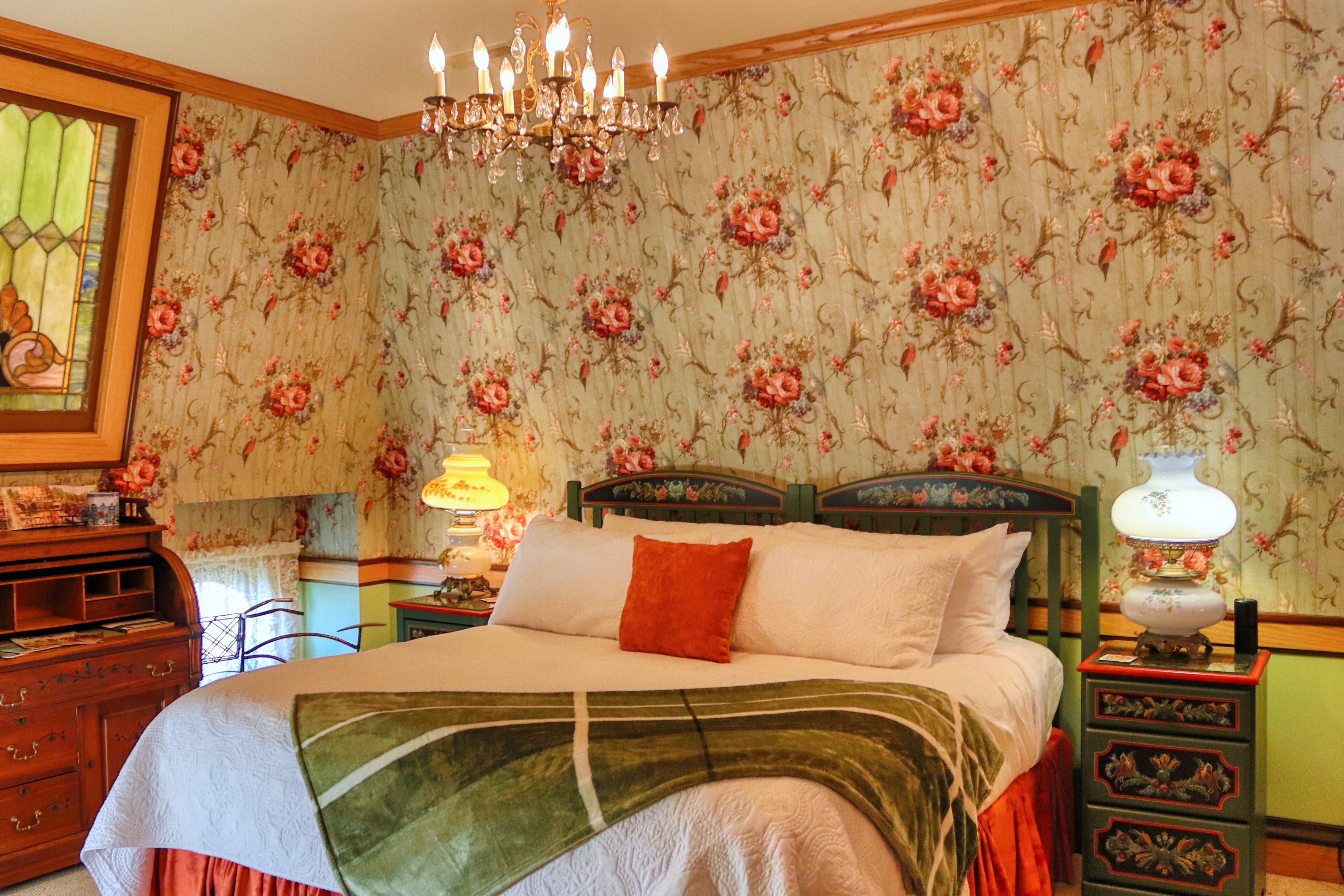 Vintage-style bedroom with floral wallpaper, a bed with a red skirt, wooden furniture, and a chandelier.