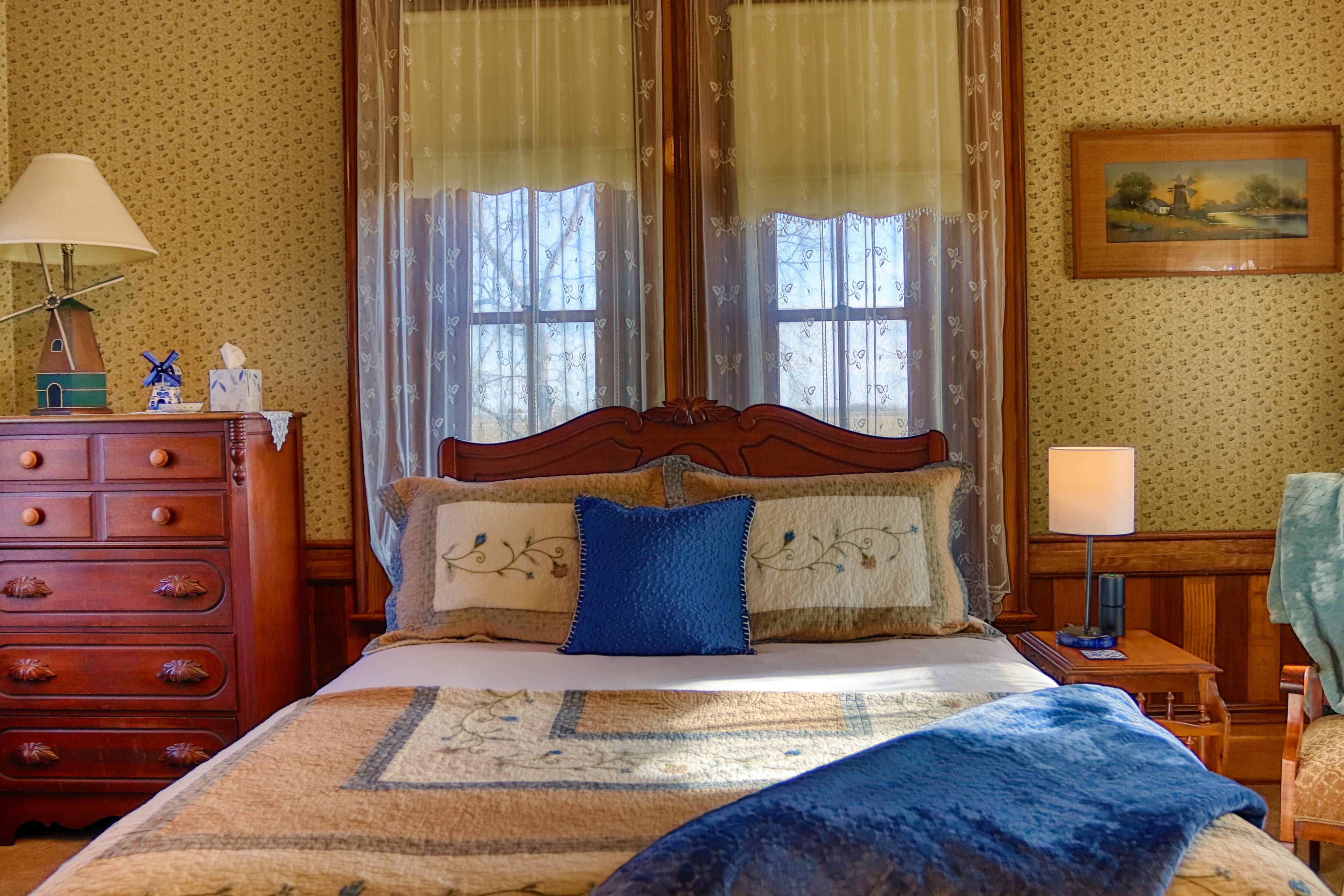 Traditional queen bedroom with wooden furniture and lace-curtained windows.