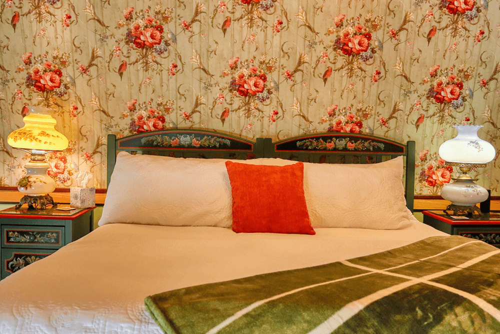A cozy bedroom featuring a floral-patterned wallpaper, a bed with decorative pillows, and vintage lamps.