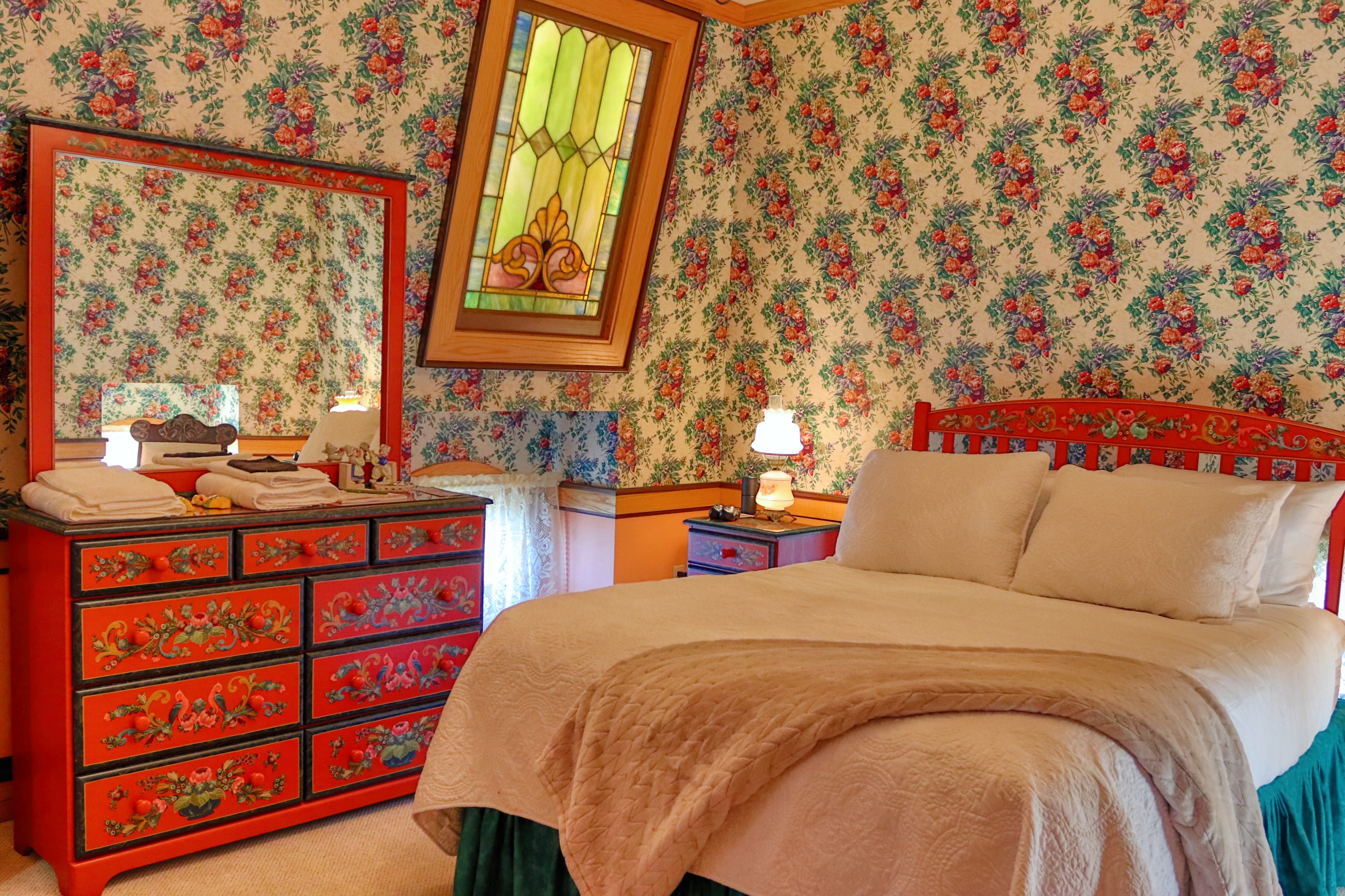 A vintage-style bedroom with custom Pella circle windows on 3 sides, floral wallpaper, hand-painted wooden furniture, air purifier, small fan, TV, a chandelier, and more!