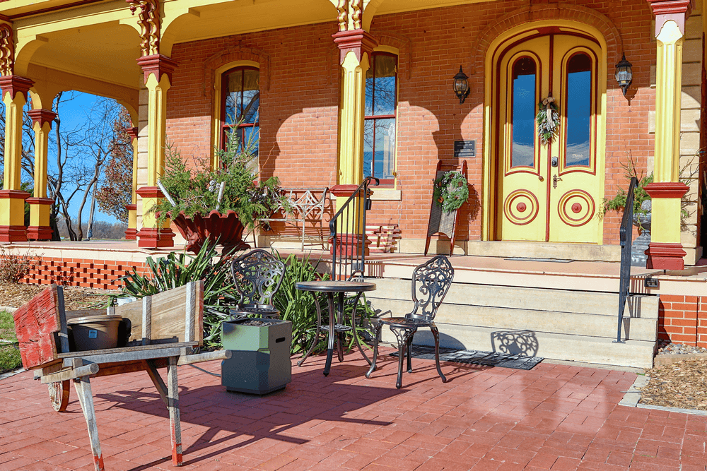 A colorful historic porch features a wheelbarrow, potted plants, and decorative chairs.